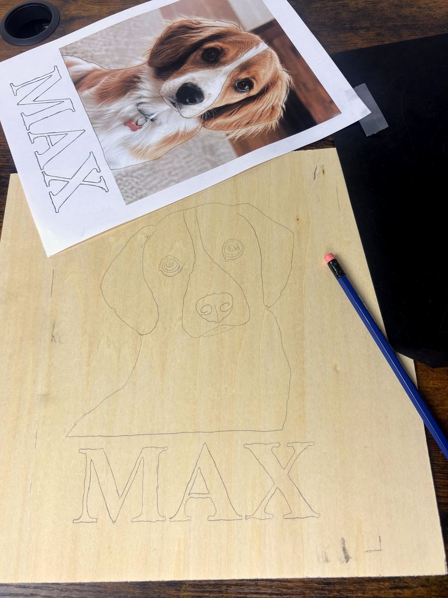 A dog's photo clipped beside a wood board with graphite transfer paper peeled back and the portrait mapped in pencil